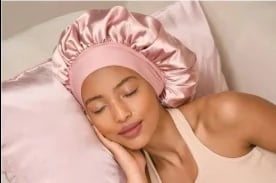 Woman wearing a pink silk bonnet, lying on pink bedding with a relaxed expression